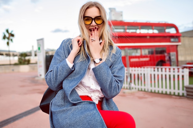 Free photo young pretty blonde woman posing on the street , wearing trendy coat and backpack, hipster student style, having fun and smiling, cute positive face.