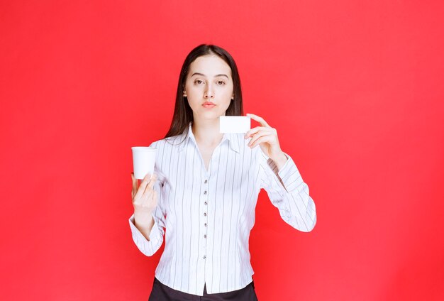 Young pretty businesswoman holding plastic cup and business card on red background. 