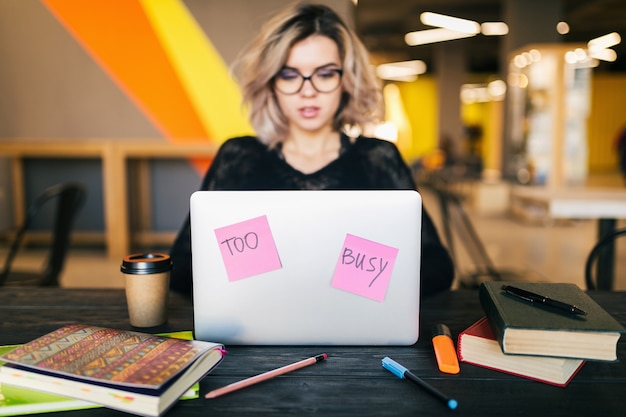 Young pretty busy woman sitting at table working on laptop in co-working office, paper stickers, wearing glasses, consentration, student in class room