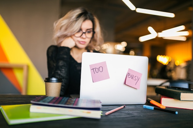 Young pretty busy woman sitting at table working on laptop in co-working office, paper stickers, wearing glasses, consentration, student in class room