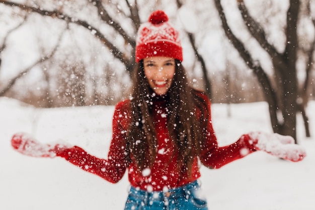 Young pretty candid smiling happy woman in red mittens and hat wearing knitted sweater walking playing in park in snow, warm clothes, having fun