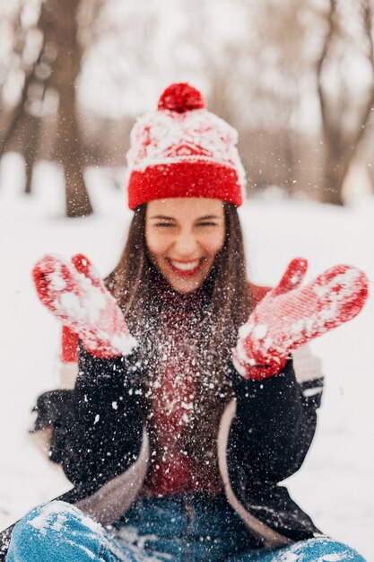 Young pretty candid smiling happy woman in red mittens and knitted hat wearing black coat walking playing in park in snow, warm clothes, having fun