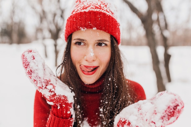 Young pretty candid smiling happy woman with funny face expression in red mittens and hat wearing knitted sweater walking playing in park in snow, warm clothes, having fun