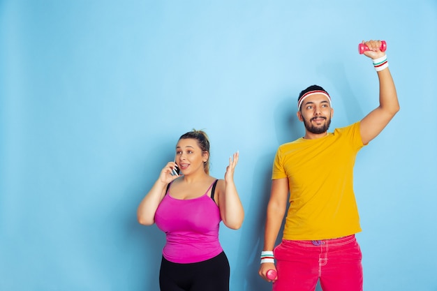 Free photo young pretty caucasian couple in bright clothes training on blue background concept of sport, human emotions, expression, healthy lifestyle, relation, family. he's training, she's talking on phone.