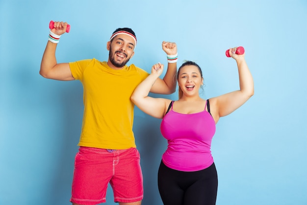 Young pretty caucasian couple in bright clothes training on blue background Concept of sport, human emotions, expression, healthy lifestyle, relation, family. Training with weights, have fun.