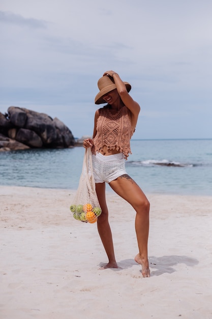 Young pretty caucasian tanned fit woman in knitted clothes on tropical beach wearing straw hat holding ecology reusable string bag full of fruits apple orange grapes