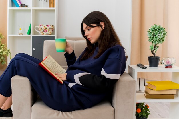 Young pretty caucasian woman sitting on armchair in designed living room holding cup with book on legs touching and reading book