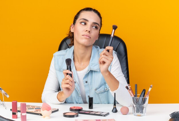 Young pretty caucasian woman sitting at table with makeup tools holding and looking at makeup brushes 