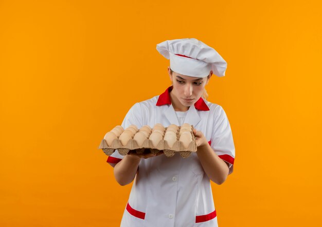 Young pretty cook in chef uniform holding and looking at carton of eggs 