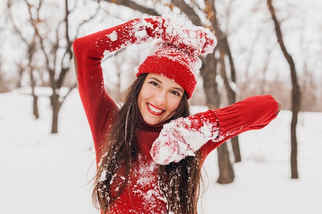 Young pretty excited candid smiling happy woman in red mittens and hat wearing knitted sweater walking playing in park in snow, warm clothes, having fun