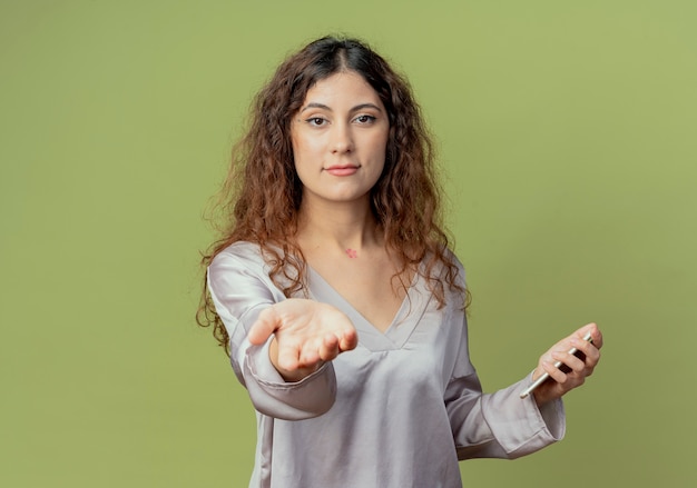 young pretty female office worker holding phone and holding out hand isolated on olive green wall