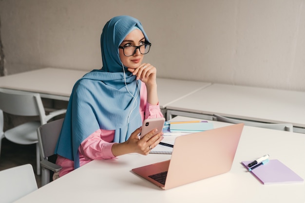 Young pretty modern muslim woman in hijab working on laptop in office room, education online