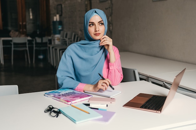 Young pretty modern muslim woman in hijab working on laptop in office room, education online