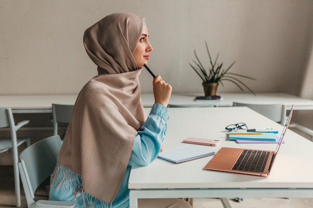 Young pretty modern muslim woman in hijab working on laptop in office room, education online