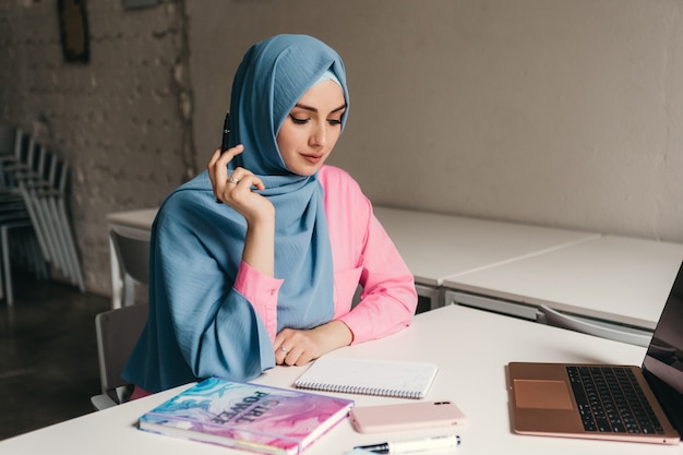 Young pretty modern muslim woman in hijab working on laptop in office room, education online