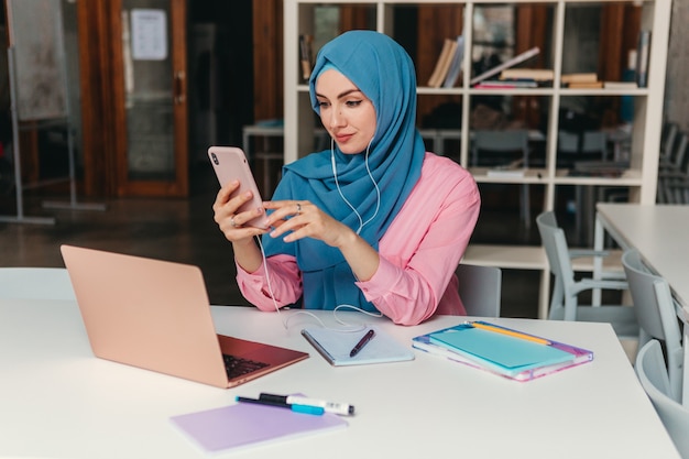 Young pretty modern muslim woman in hijab working on laptop in office room, education online