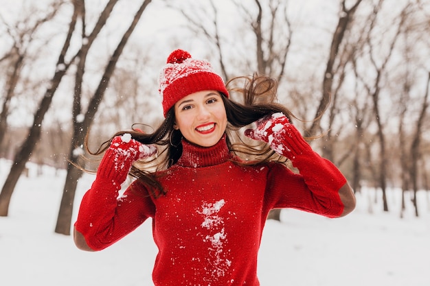 Young pretty smiling happy woman in red mittens and hat wearing knitted sweater walking in park in snow, warm clothes, having fun, waving long hair