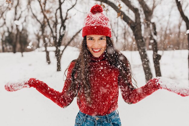 Young pretty smiling happy woman in red mittens and hat wearing knitted sweater walking in park in snow, warm clothes, having fun