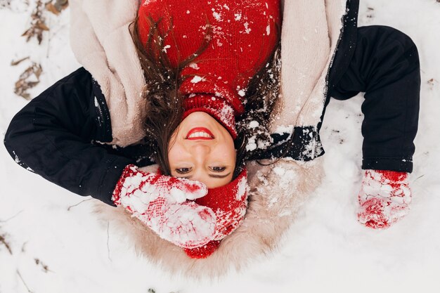 Young pretty smiling happy woman in red mittens and knitted hat wearing winter coat lying in park in snow, warm clothes, view from above