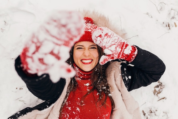 Young pretty smiling happy woman in red mittens and knitted hat wearing winter coat lying in park in snow, warm clothes, view from above