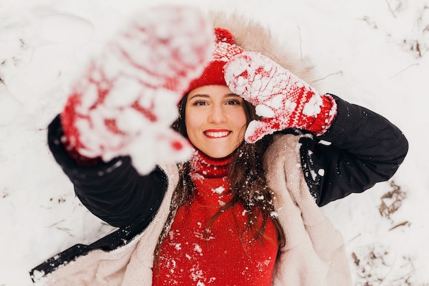 Young pretty smiling happy woman in red mittens and knitted hat wearing winter coat lying in park in snow, warm clothes, view from above