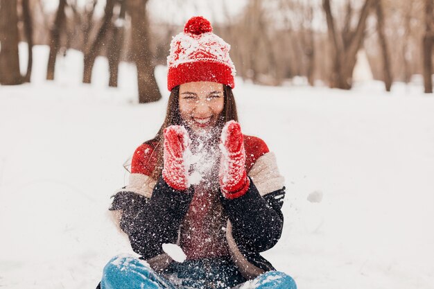 Free Photo young pretty smiling happy woman in red mittens and knitted hat wearing winter coat sitting on snow in park, warm clothes