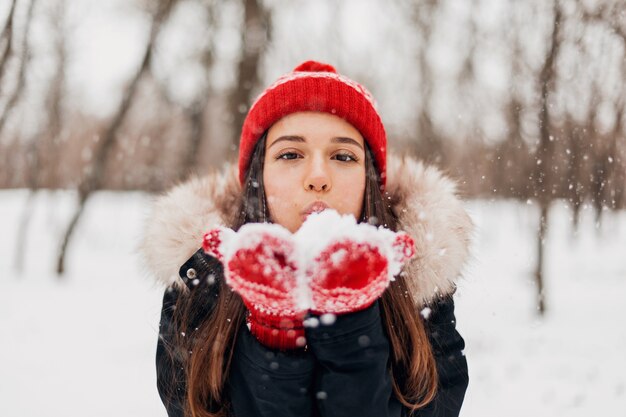 Young pretty smiling happy woman in red mittens and knitted hat wearing winter coat, walking in park, blowing snow