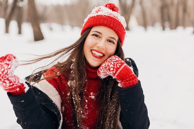 Young pretty smiling happy woman in red mittens and knitted hat wearing winter coat, walking in park, playing with snow in warm clothes