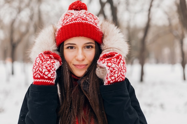 Young pretty smiling happy woman in red mittens and knitted hat wearing winter coat with fur hood, walking in park in snow, warm clothes