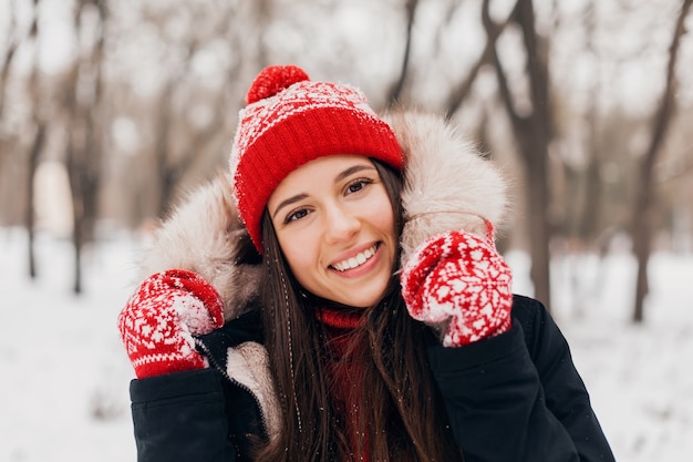 Young pretty smiling happy woman in red mittens and knitted hat wearing winter coat with fur hood, walking in park in snow, warm clothes