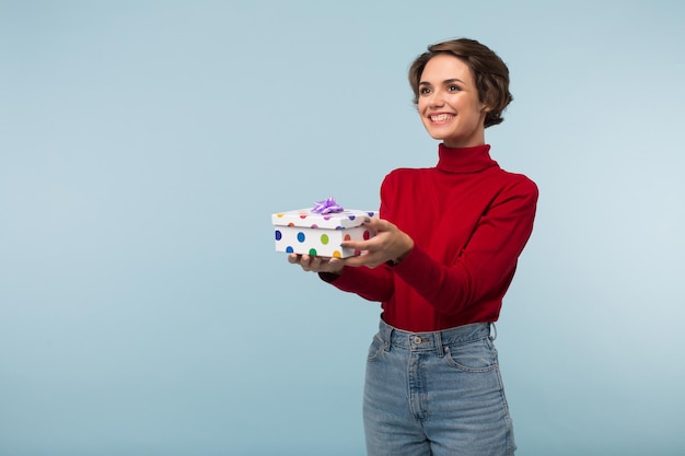 Young pretty smiling woman with dark short hair in red sweater holding little present box in hands while happily looking aside over blue background