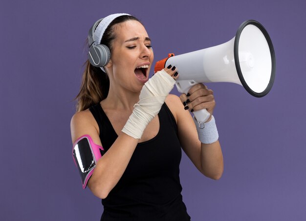 Young pretty sporty girl wearing headband wristbands headphones and phone armband with injured wrist wrapped with bandage talking by speaker isolated on purple wall