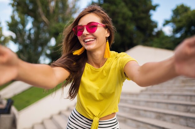 Young pretty stylish smiling woman making selfie in city park, positive, emotional, wearing yellow top, pink sunglasses, summer style fashion trend, long hair, having fun