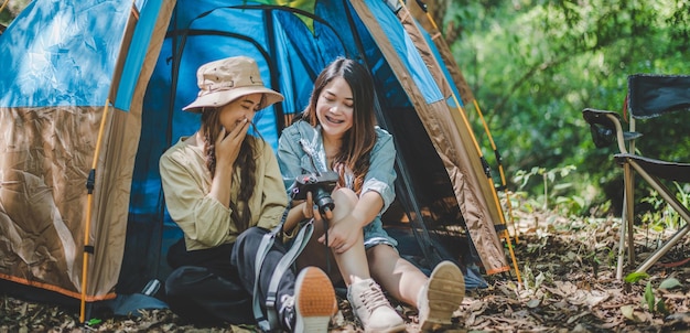 Free photo young pretty taking photo by camera and showing picture to her friend while sitting at the camping tent in forest young asian group women travel outdoor camping