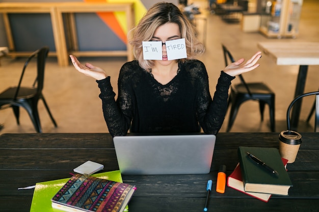 Free photo young pretty tired woman with paper stickers on glasses sitting at table in black shirt working on laptop