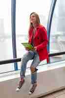 Free photo young pretty woman in casual outfit holding tablet laptop in urban building, wearing jeans, pink trench jacket, sitting at window with city view