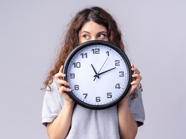 Young pretty woman holding clock looking at side from behind it isolated on white wall