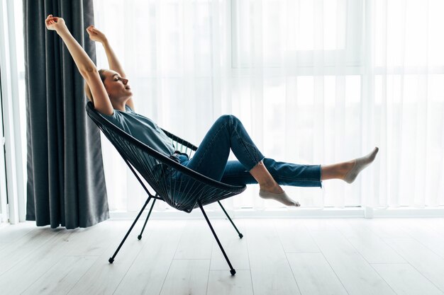 Young pretty woman at home sitting on modern chair in front of window relaxing in her living room