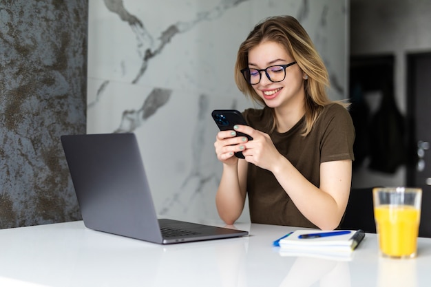 Free Photo young pretty woman sits at the kitchen table using a laptop and use on a cell phone