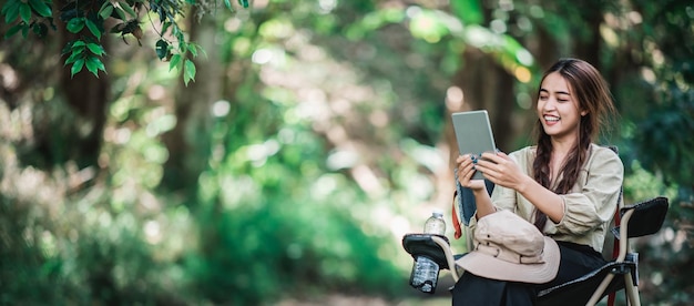 Free photo young pretty woman sitting in chair and use tablet video call while camping in nature park copy space