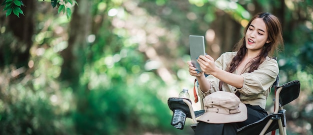 Young pretty woman sitting in chair and use tablet video call while camping in nature park copy space