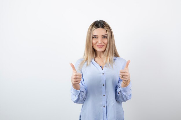 young pretty woman standing and giving thumbs up on white wall.