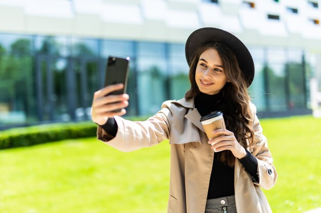 Young pretty woman student wear in coat and hat holding coffee to go take selfie on the phone in the street