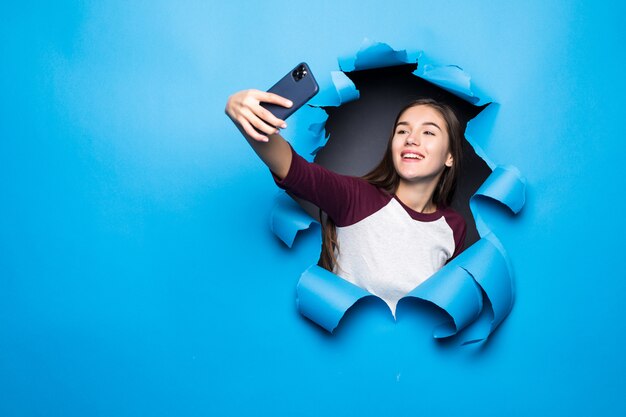 Young pretty woman take selfie on the phone while looking through blue hole in paper wall.