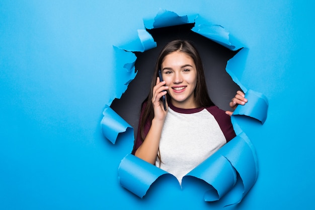 Young pretty woman talk phone while looking through blue hole in paper wall.