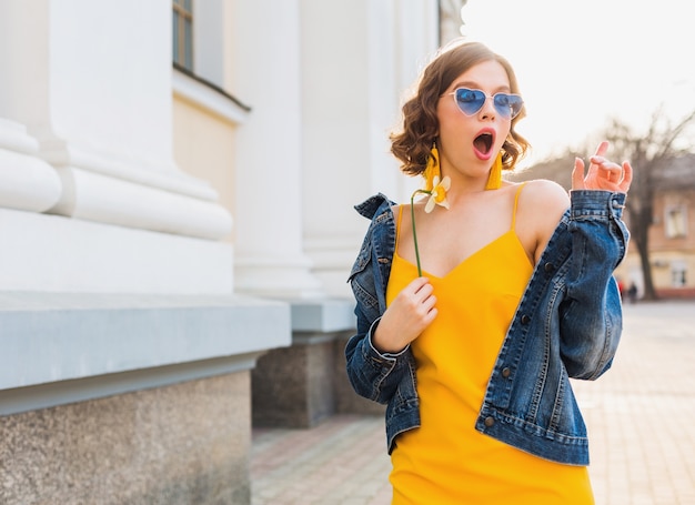 Free photo young pretty woman with surprised face expresssion, emotional, shocked emotion, wearing stylish apparel, denim jacket, yellow top, holding flower, sunny summer, trendy funny sunglasses