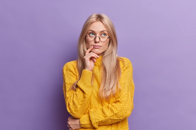 Free Photo young pretty young woman thinks of ideas concentrated above stands thoughtful and keeps hand on face stands in thoughtful pose wears round glasses yellow sweater 