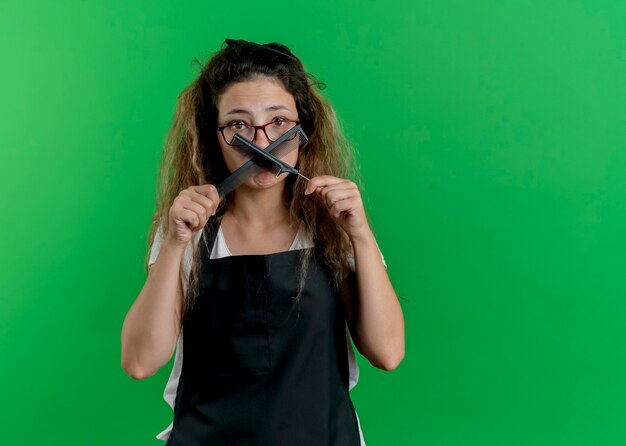 Young professional hairdresser woman in apron holding hair combs at camera confused with sad expression 