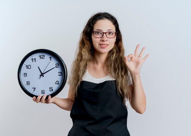 Young professional hairdresser woman in apron holding wall clock looking at front showing ok sign standing over white wall