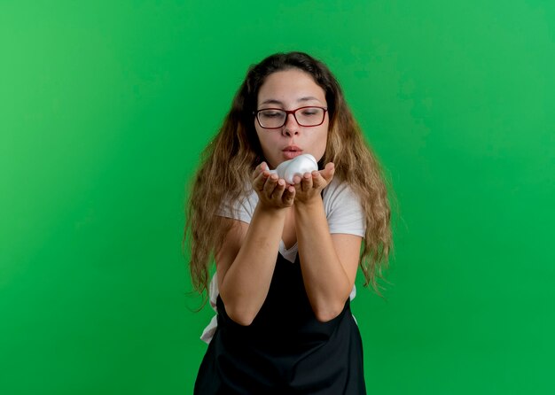 Young professional hairdresser woman in apron with shaving foam in hands blowing on it standing over green wall